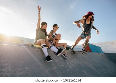 Two Young Women Sitting On Skating Ramp With A Girl Practising Skateboarding. Skateboarding Woman Riding Skateboard At Skate Park Ramp With Friends Sitting By.