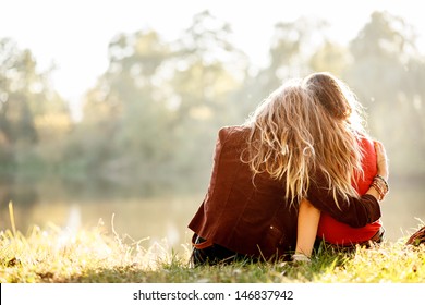 two young women sitting on grass hugging rear view
