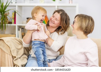 Two Young Women Are Sitting On The Couch By The Seyu At Home In The Living Room And Playing With Their Little Daughter. Gay Lesbian Couple At Home With A Baby. Lesbian Family 