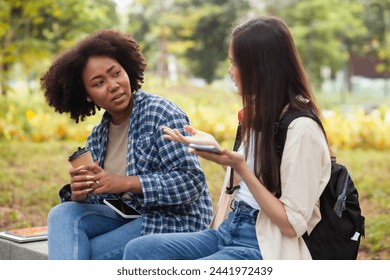 Two young women sitting on a bench in a park, one of them holding a cell phone. Scene is casual and relaxed, as the women are enjoying each other's company and having a conversation - Powered by Shutterstock