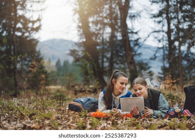 Two young women sitting on the ground in a forest, looking at a tablet together. They are surrounded by nature, with trees and mountains in the background. Both are smiling and engaged with the device - Powered by Shutterstock