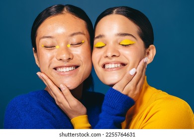 Two young women smile and embrace each other with makeup on their faces. Female friends in their 20’s feeling self-assured as they embrace the gen z beauty trends of graphic eyeliner and eyeshadow. - Shutterstock ID 2255764631