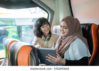 two young women smiling while chatting and using tablets sit on the bus - Powered by Shutterstock