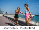 Two young women stretching legs before running on beachfront promenade in barcelona