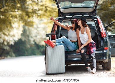 Two Young Women With Suitcases On Car Trip.They Are Sitting In Car Back And Resting After Long Ride.Taking Selfie.