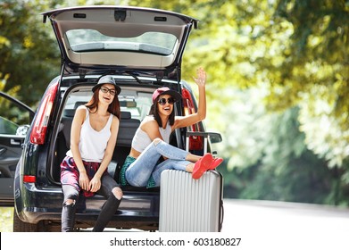 Two Young Women With Suitcases On Car Trip. They Are Sitting In Back Of The Car, Resting After Long Ride And Having Fun.