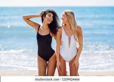 Two young women in swimsuits on a tropical beach. Funny caucasian and arabic females wearing black and white swimwear kneeling on the beach.