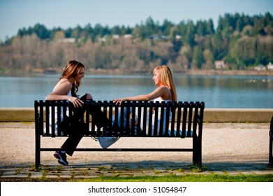 Two Young Women Talking On A Bench By A Lake