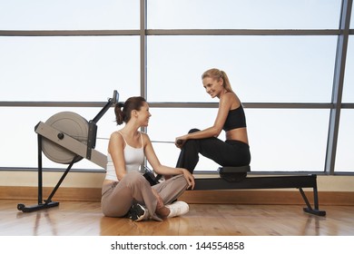 Two Young Women Talking At Rowing Machine In Health Club