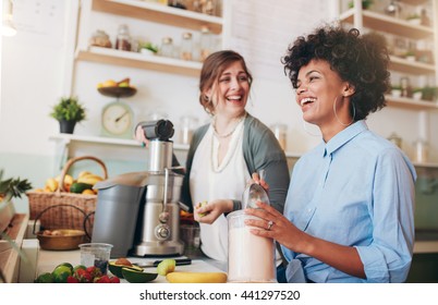 Two Young Women Talking And Smiling While Working At Bar Counter. They Are Preparing Fruit Juice.