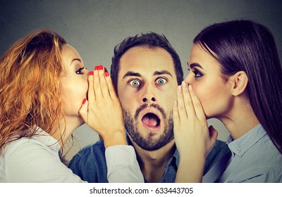 Two young women telling whispering secret gossip in the ear to an amazed shocked man with wide open mouth. Human emotions face expression reaction. Hot news concept 
