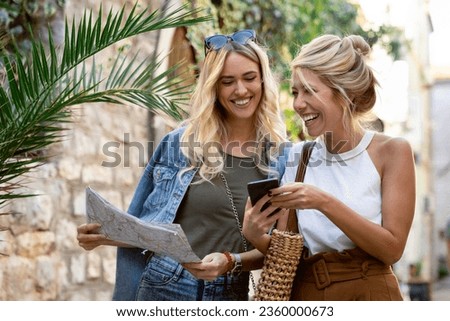 Two young women tourists using mobile phone in the city. Happy blonde sisters walking in the cobbled street and having fun on vacation.