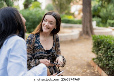 Two young women are using their smartphones and smiling while sitting on a bench in a park, enjoying a moment of relaxation and connection together - Powered by Shutterstock