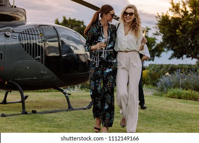 Two Young Women Walking Away From A Helicopter With A Glass Of Wine In Their Hands. Best Friends Walking Together With Wine And Having Fun.