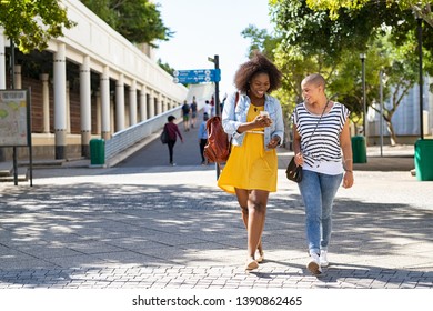 Two Young Women Walking While Using Mobile Phone On Urban City Street. Cheerful Woman Doing Online Shopping With Friend Outdoor. African American Girl Showing Smartphone Screen To Friend.