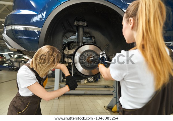 Two young women working in
autoservice, repairing brake disc of car. Professional female
mechanics in black gloves using tools, fixing automobile lifted on
bridge.