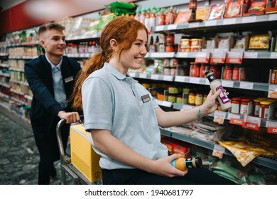 Two Young Workers Having Fun While Working In A Supermarket. Woman Sitting On Cart Stocking Shelves With Man Colleague Pushing In Supermarket Aisle.