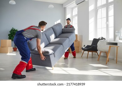 Two Young Workers Lift Up Heavy Sofa Together. Young Men From Moving Company And Lorry Delivery Service Removing Things From The House, Carrying Furniture And Other Belongings