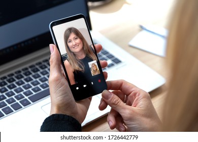 Two Youngs Woman Talking To Each Other Through A Video Call On A Smartphone. Blonde Woman Having A Video Chat With Other Girl On Mobile Phone. Woman Working In The Office Or Home, In Front At Laptop.