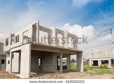 A two-story concrete house under construction, featuring an unfinished structure with open window spaces, set on a dirt lot with a clear sky and nearby building sites in the background.