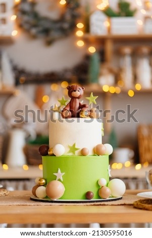 two-tiered white-green cake with balls, stars and a teddy bear on the kitchen table with electric garland for a children's holiday. the tradition of baking a birthday cake. professional confectioner.