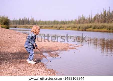a two-year-old child in a denim suit plays outdoors near water. A blond-haired little girl stands on the bank of the river and sticks muddy water with a stick. nonstop laid-back lifestyle photography