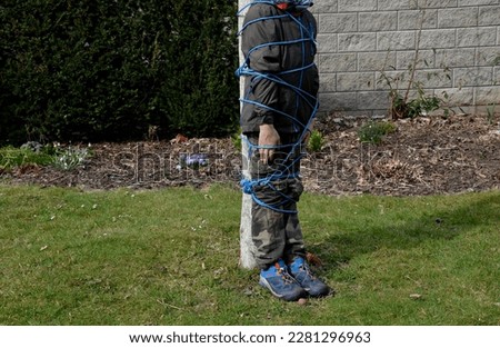 tying with blue plastic rope to the tree. innocent children's game of pirates, or school bullying. a friend stands by a tree trunk and is wrapped in a rope. hands along the body, abusing the boy
