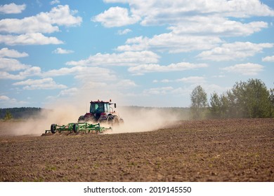 Typical agricultural scene tractor cultivation in field in clouds of dust drives off into the distance with birches. Selective focus red farm tractor with green plow under blue sky with copyspace.