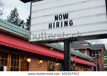 Typical american now hiring sign in black letters over the white background with local small business building 