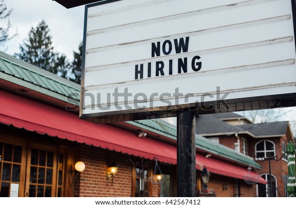 Typical american now\
hiring sign in black letters over the white background with local\
small business building\
