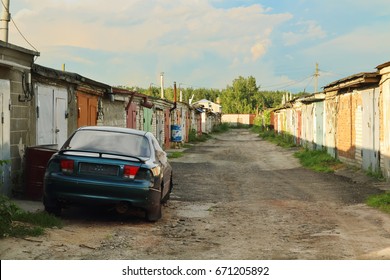 Typical Complex Of Old Concrete Garages With Closed Metallic Doors In Russia. Driveway Road Through Garages With Perspective And Old Blue Parked Car In Summer Day.