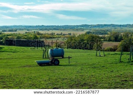 Typical French rural landscape in the village of Leyment, Auvergne-Rhône-Alpes, France. Green field, blue watertank, trees and hills in the horizon, evening light and a pile of firewood.