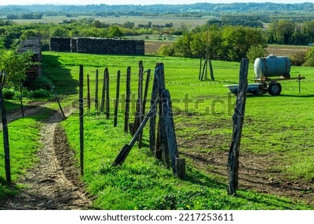 Typical French rural landscape in the village of Leyment, Auvergne-Rhône-Alpes, France. Green field, blue watertank, trees and hills in the horizon, wood fence and a pile of firewood.