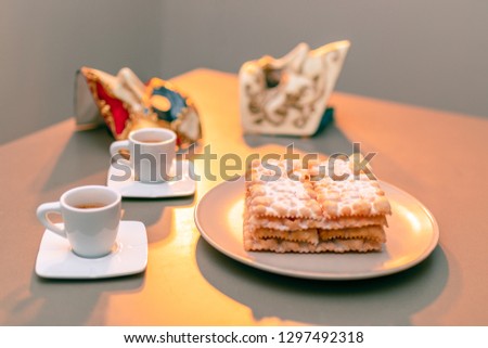 Typical Italian carnival fritters (Chiacchiere di Carnevale) dusted with powdered. Composition with two cups of coffee and in the background are Venetian carnival masks. Homemade Italian sweets.