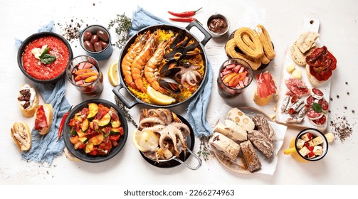 Typical spanish tapas concept. Concept include jamon, chorizo sausage, brushettas, bowl with olives, shrimp, pan with paella, cheese, sangria, churros on a white background. Top view.