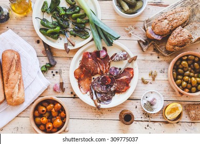 Typical spanish tapas concept. Concept include variety slices smoked cut dry sausages, ham, bowls with olives, small bbq peppers, bread baguette, veggies from above on wooden table. Rustic style. - Powered by Shutterstock