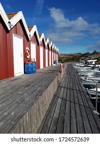 Typical Swedish red sunlit wooden boat huts against a blue sky with a few clouds on the island of Hönö with a wooden jetty with partially visible boats at the harbor.