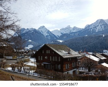 A Typical Swiss Chalet Style House In A Village Surrounded By Snowy Pine Trees And Mountains.