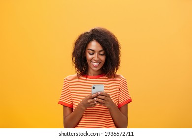 Typing answer to party invitation via smartphone message app holding device over chest smiling joyfully at gadget screen, texting or playing funny game, waste time in subway over orange wall