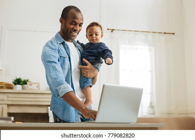 Typing. Attractive alert young afro-american father holding his little son and typing on the laptop while standing at the table and a fireplace in the background - Powered by Shutterstock