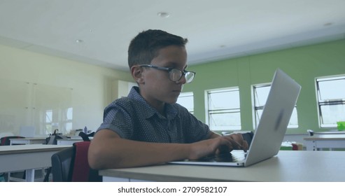 Typing boy wearing glasses and patterned shirt at classroom desk, using open silver laptop. Student, spectacles, school, learning, technology, device, sunlit