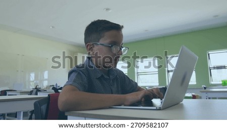 Typing boy wearing glasses and patterned shirt at classroom desk, using open silver laptop. Student, spectacles, school, learning, technology, device, sunlit