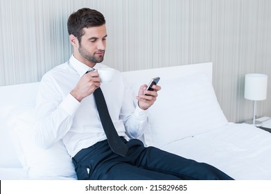 Typing Business Message. Confident Young Man In Shirt And Tie Drinking Coffee And Holding Mobile Phone While Lying In Bed At The Hotel Room 