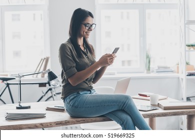 Typing business message. Confident young woman in smart casual wear holding smart phone and looking at it with smile while standing near her working place