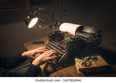 Typing hands of a writer looking for inspiration to start a new novel on his vintage typewriter in a retro style studio: a wooden desk lit by a lamp, an old book and a pair of glasses.