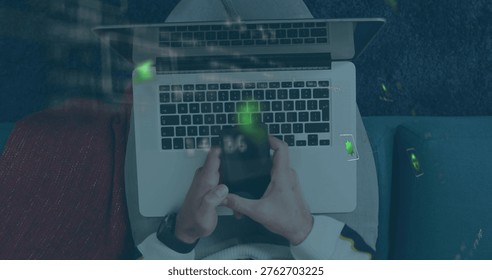Typing man in light sweater using laptop on lap on sofa, holding smartphone reflecting green. Remotework, computer, cozy, cushions, lounge, topdown, reflection