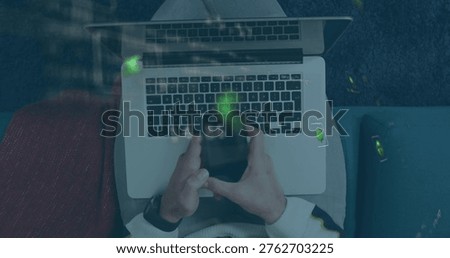 Typing man in light sweater using laptop on lap on sofa, holding smartphone reflecting green. Remotework, computer, cozy, cushions, lounge, topdown, reflection