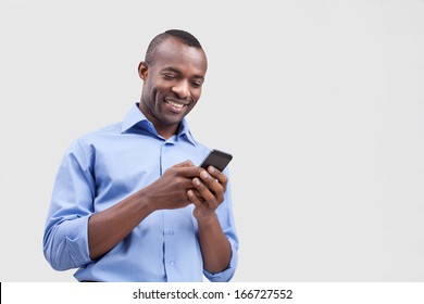 Typing A Message. Cheerful Black Man Typing Something On The Mobile Phone And Smiling While Standing Isolated On Grey