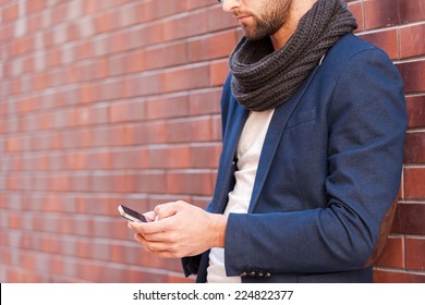 Typing message to friend. Side view of handsome young man in smart casual wear holding mobile phone while leaning at the brick wall
