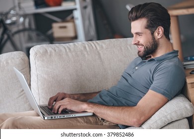 Typing New Blog Post. Side View Of Handsome Young Man Using His Laptop With Smile While Sitting On The Couch At Home